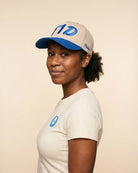 A woman wearing a cream-colored t-shirt and a matching cream and blue baseball cap, photographed against a solid light beige background.