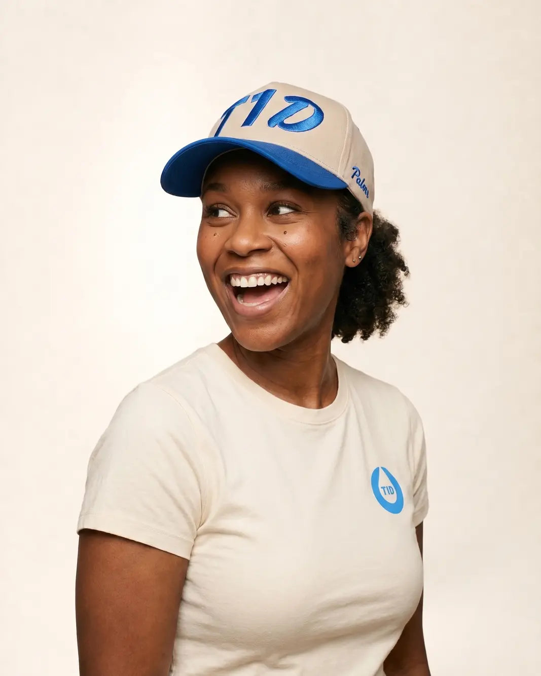 A smiling woman wearing a beige and blue cap with a blue logo and a matching beige t-shirt with a blue logo, photographed against a plain light beige background.