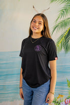 Person wearing a black t-shirt with a logo, standing in front of a scenic background with water and plants.
