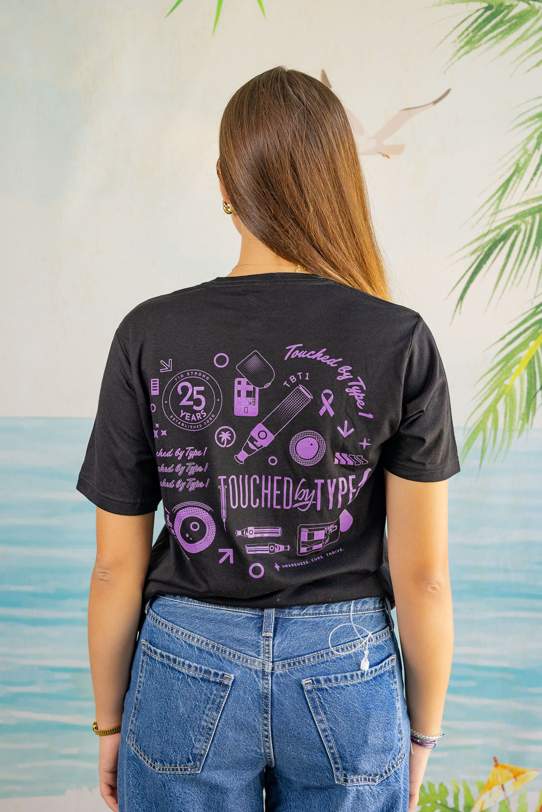 Person wearing a black t-shirt with purple graphics and text, standing against a light-colored wall with greenery.