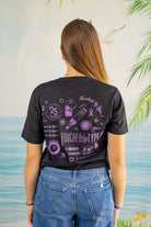 Person wearing a black t-shirt with purple graphics and text, standing against a light-colored wall with greenery.