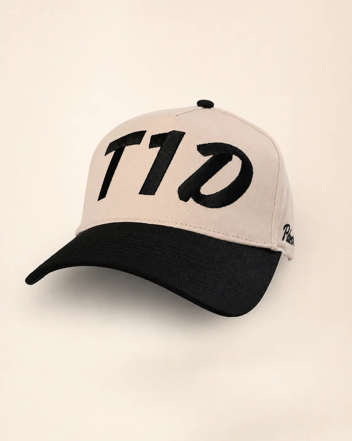 A baseball cap with a light beige crown and a black curved brim, featuring a large black stylized 'T1D' logo on the front and a small black script logo on the side, set against a plain beige background.