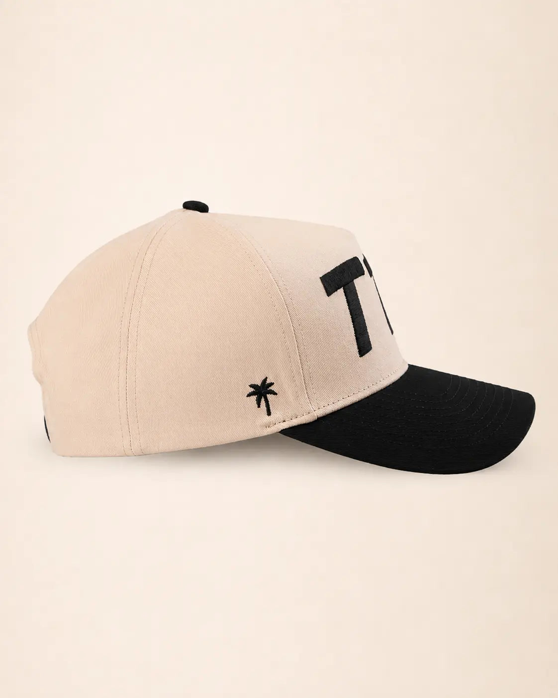 A two-tone baseball cap with a light beige crown and a black curved brim, featuring a black embroidered palm tree logo and a large black letter 'T1D' on the side, set against a solid cream background.