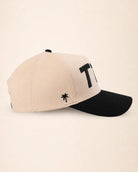A two-tone baseball cap with a light beige crown and a black curved brim, featuring a black embroidered palm tree logo and a large black letter 'T1D' on the side, set against a solid cream background.