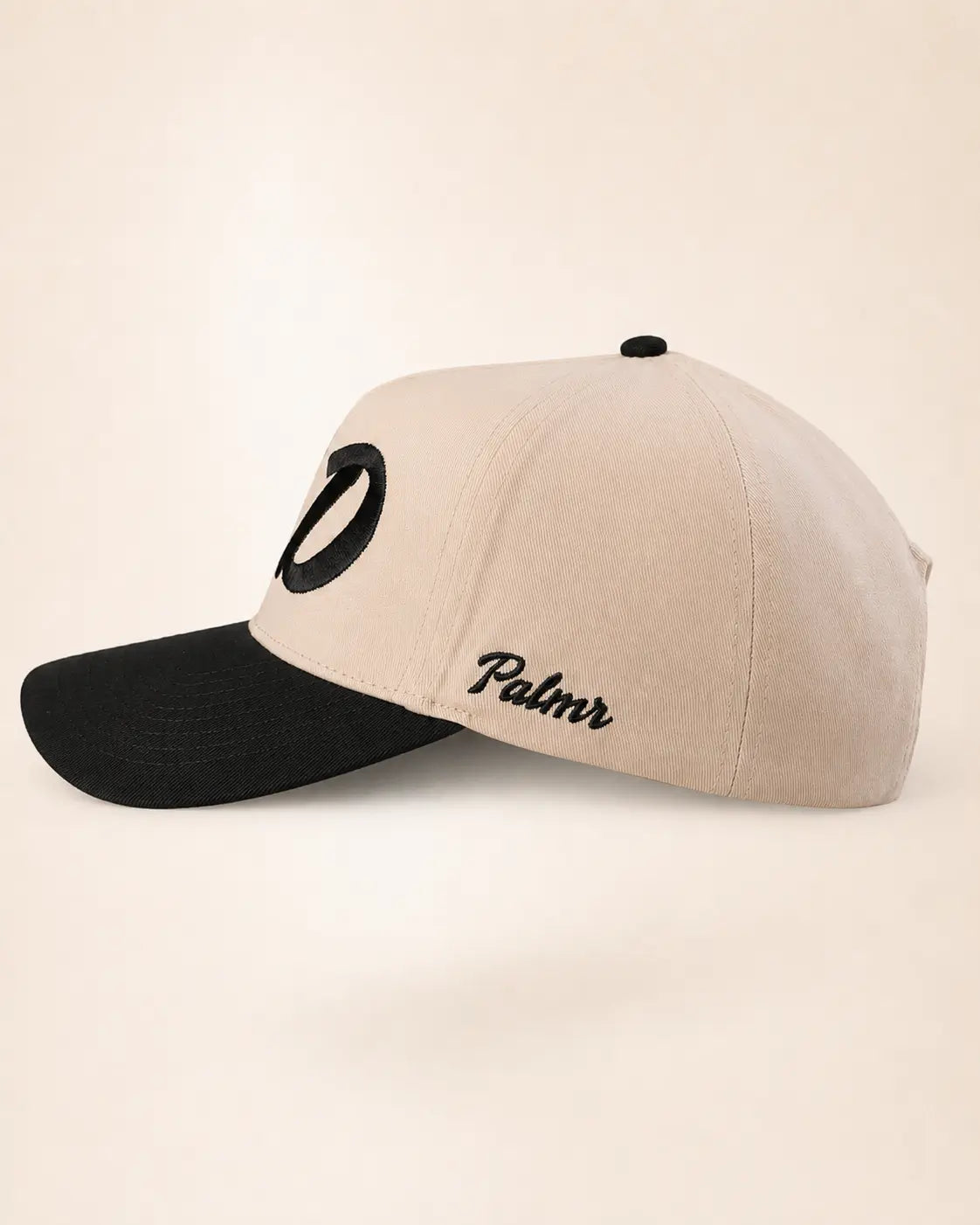 A baseball cap with a beige crown and a black brim, featuring a black embroidered oval logo on the front and the word 'Palmr' in black script on the side, set against a plain beige background.