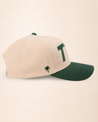 A structured baseball cap in cream and dark green, featuring a large green embroidered letter 'T1d' on the side and a small green palm tree logo on the front, set against a plain cream background.