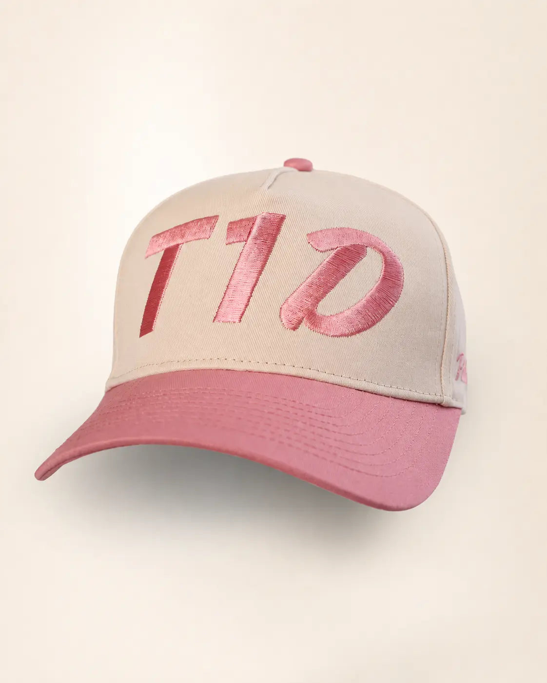 A structured baseball cap with a light beige crown and a dusty pink brim, featuring large metallic pink embroidered letters on the front.