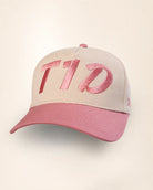 A structured baseball cap with a light beige crown and a dusty pink brim, featuring large metallic pink embroidered letters on the front.