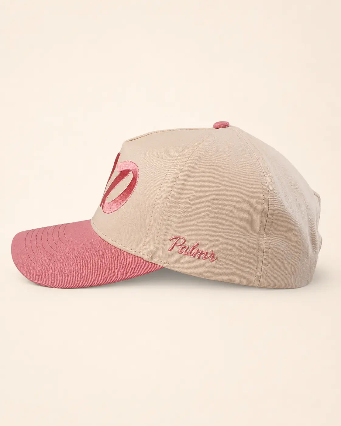 A structured baseball cap in beige with a contrasting dusty pink brim, featuring a pink ribbon embroidery on the front and 'Palmr' script embroidery on the side, set against a solid cream background.