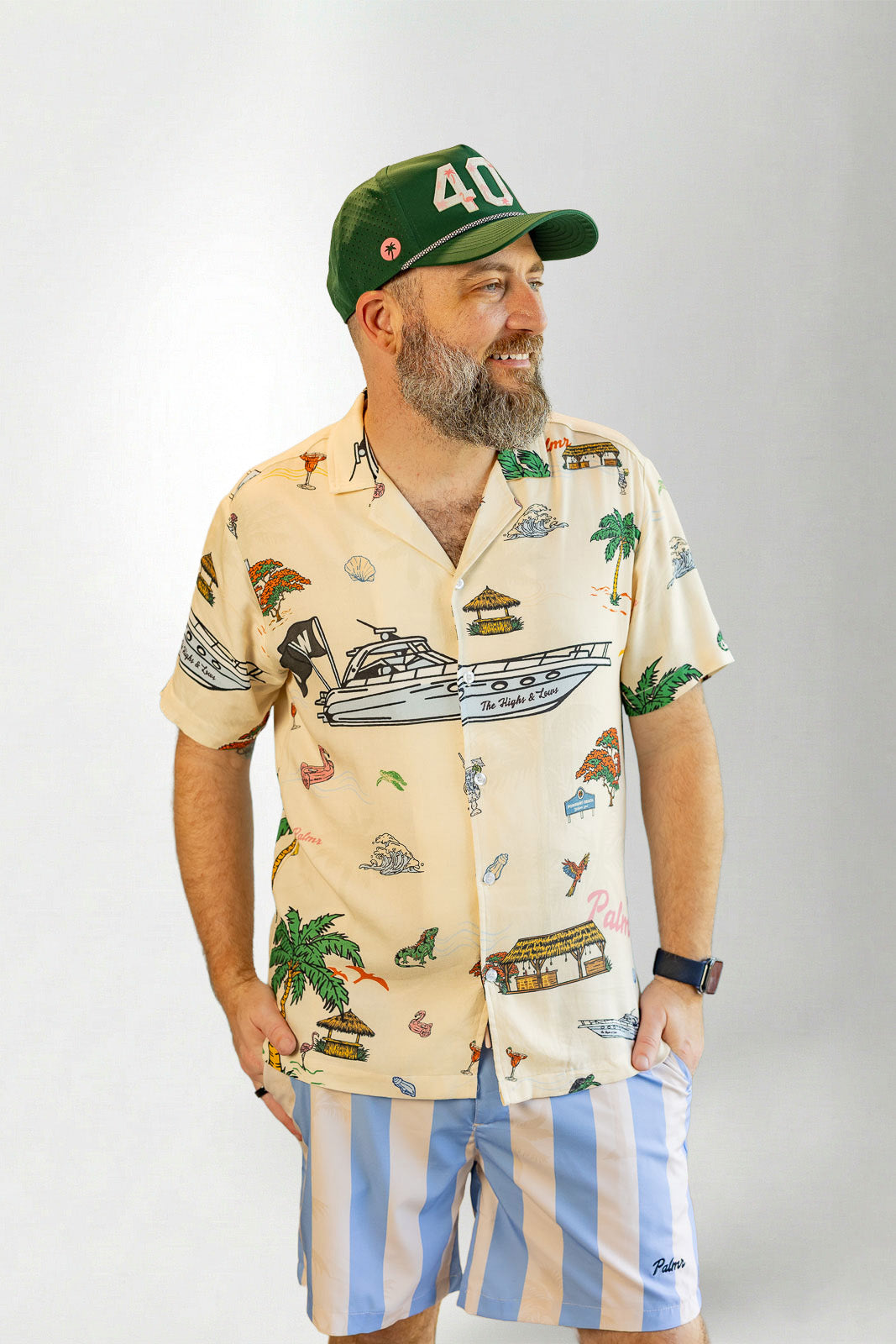 Man wearing a patterned shirt and striped shorts with a cap on a plain background