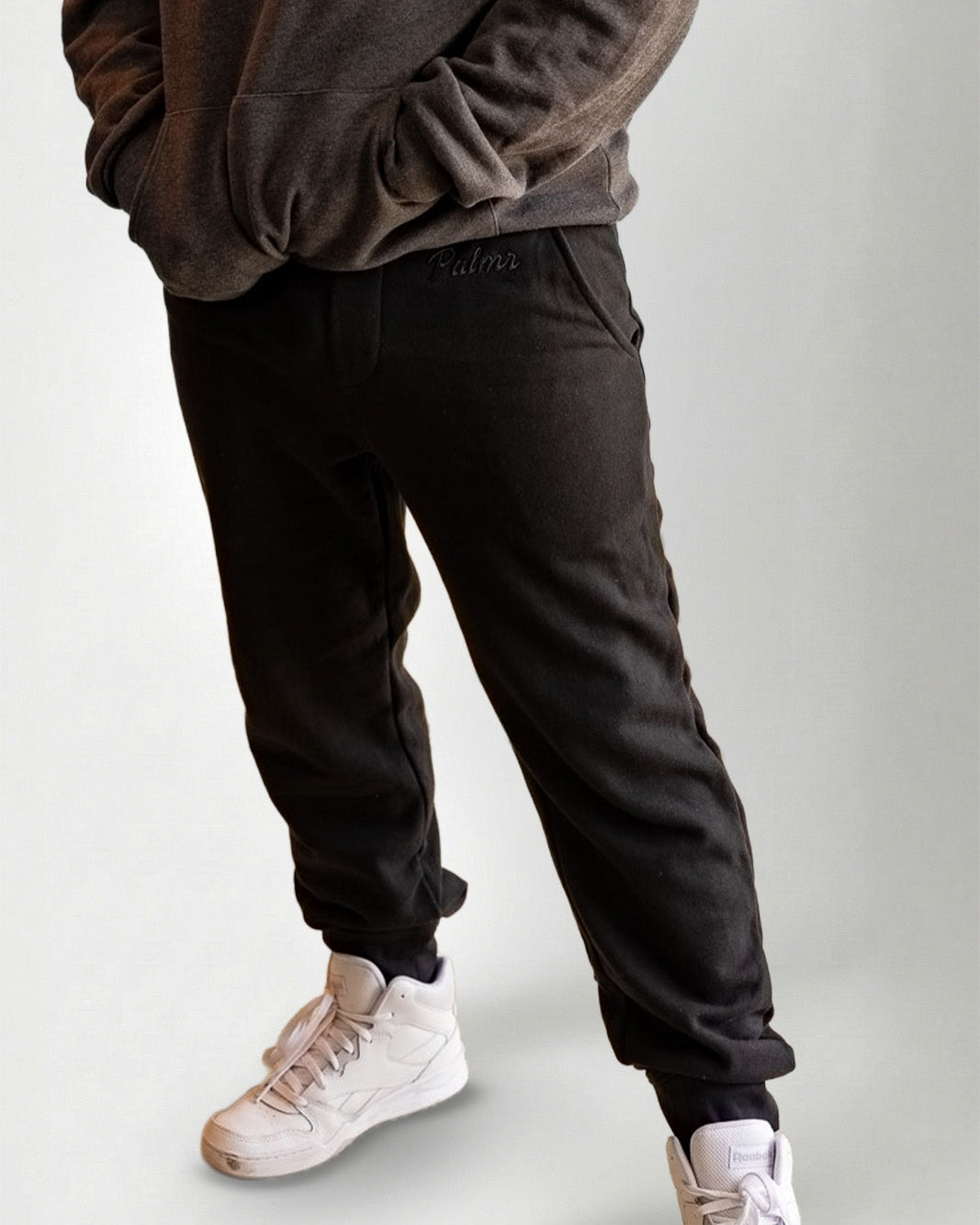 Person wearing a dark gray hoodie and pants with white sneakers on a light gray background