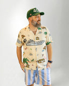 Man wearing a patterned shirt and striped shorts with a cap on a plain background