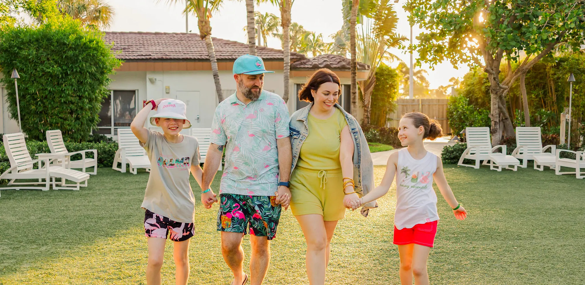 Family of four walking together in a park-like setting with palm trees and a house in the background.