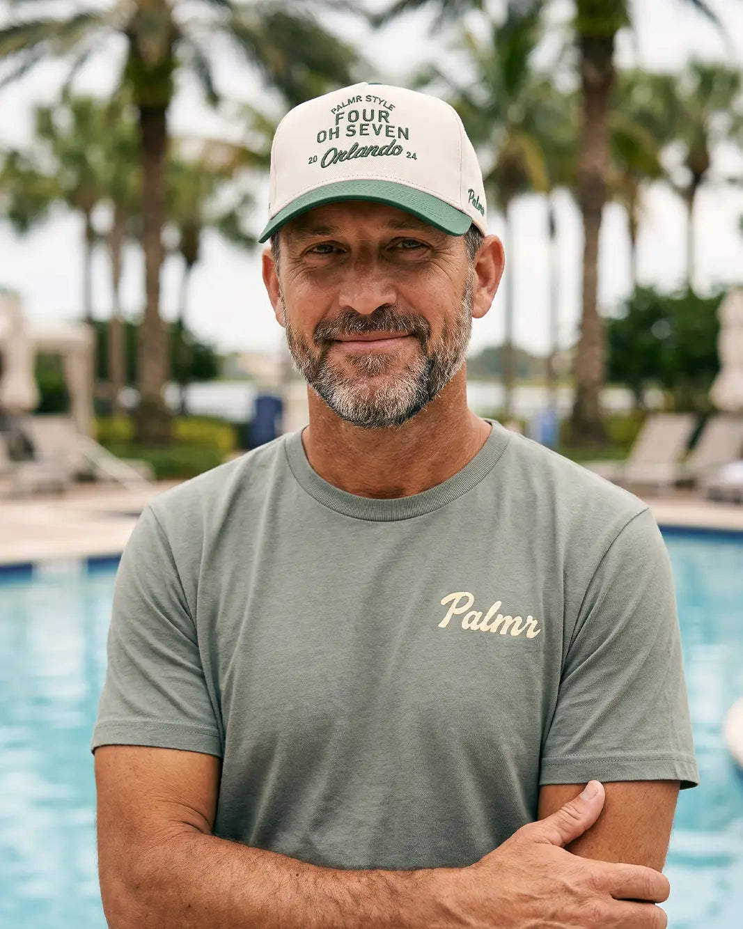 A man standing by a swimming pool wearing a light olive green t-shirt with 'Palmr' printed in gold on the chest, and a cream baseball cap with green trim and text.