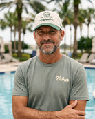 A man standing by a swimming pool wearing a light olive green t-shirt with 'Palmr' printed in gold on the chest, and a cream baseball cap with green trim and text.