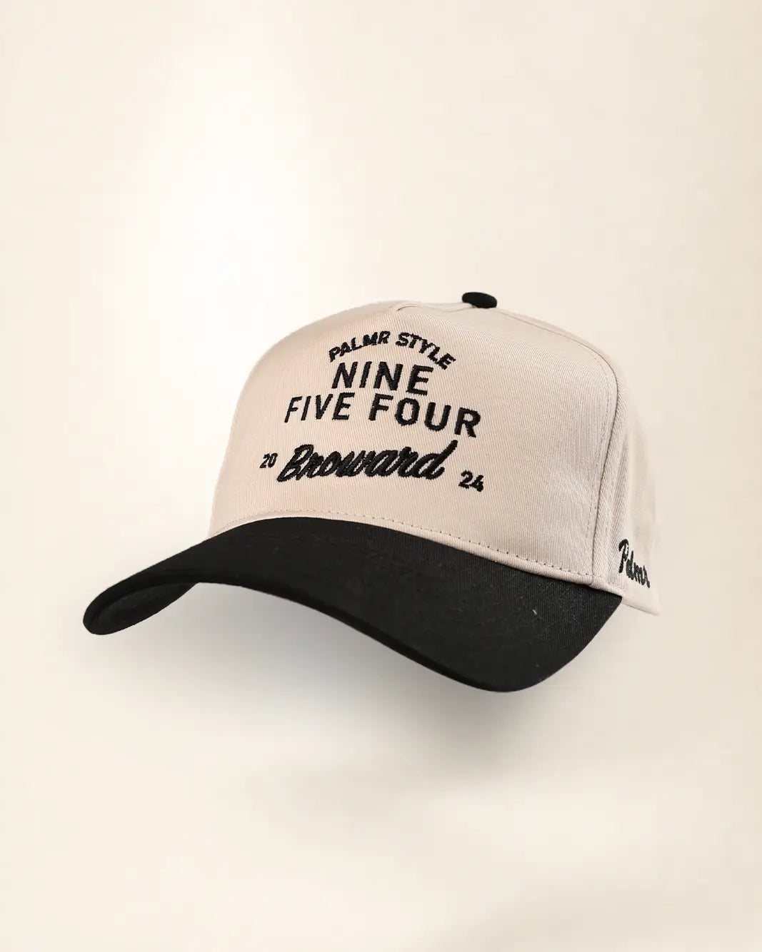A baseball cap with a cream-colored crown and a black brim, featuring black text on the front that reads 'PALMR STYLE NINE FIVE FOUR 20 Broward 24'.