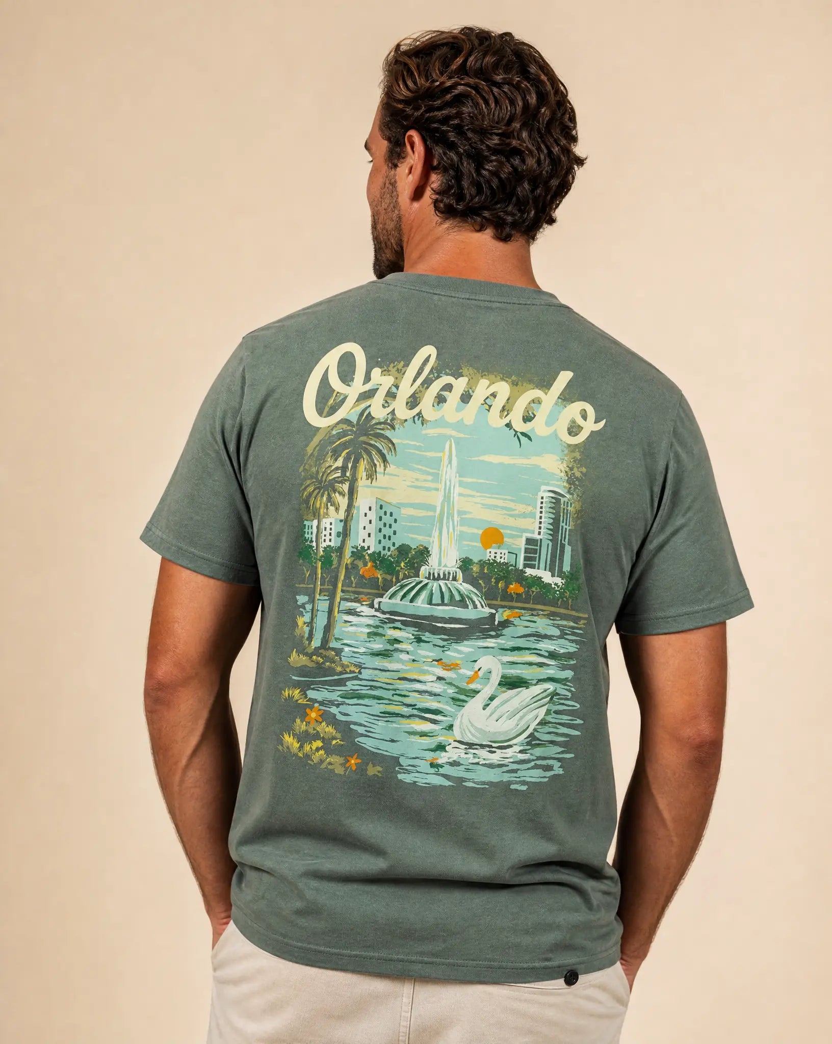 A man viewed from behind wearing a slate green t-shirt with a large back graphic depicting the Orlando skyline, fountain, palm trees, and a swan, set against a solid beige background.