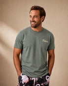 A smiling man wearing a muted sage green crewneck t-shirt with 'Palm' printed in gold script on the left chest, paired with black and pink tropical print shorts, set against a warm beige background.