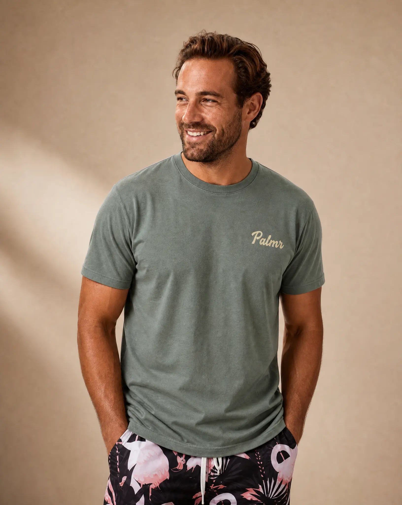 A smiling man wearing a muted sage green crewneck t-shirt with 'Palm' printed in gold script on the left chest, paired with black and pink tropical print shorts, set against a warm beige background.