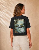 A woman shown from the back wearing a dark charcoal gray cropped t-shirt with a large graphic print of the Orlando skyline, fountain, and palm trees, paired with light beige pants against a textured beige background.