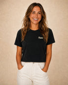 A smiling woman with long brown hair wearing a black short-sleeve crewneck t-shirt with 'Palmers' written in white script on the left chest, paired with off-white pants, set against a textured beige background.