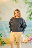 Woman wearing a gray sweatshirt with 'Frida' and beige shorts standing in front of a tropical-themed wall.