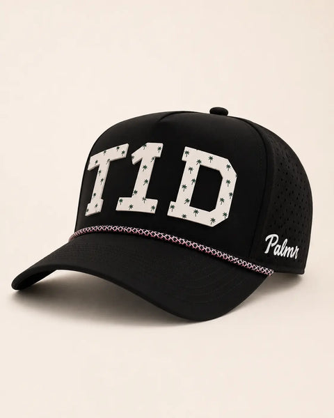 The T1D Signature Hat (Black): black rope snapback with T1D in cream palm-print letters, a pink braided rope across the brim, perforated sides.