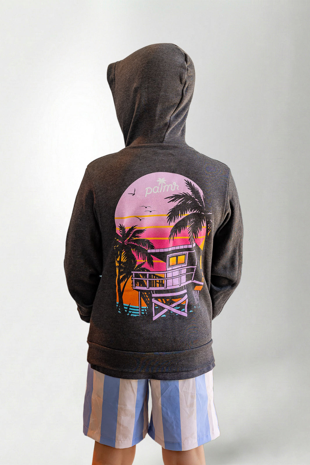 Person wearing a hoodie with a sunset and palm tree design on the back, standing against a plain background.