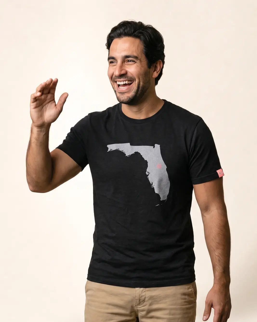 A smiling man wearing a black t-shirt with a gray Florida state map graphic on the front, paired with khaki pants, posing against a solid light beige background.