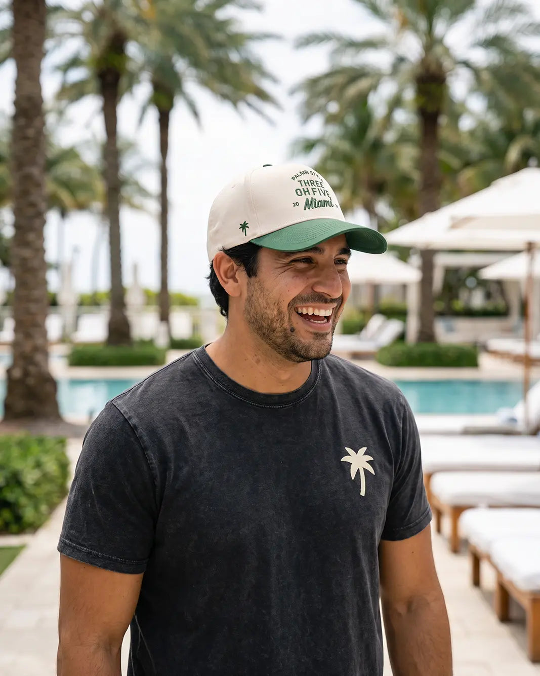 A man smiling outdoors by a pool wearing a dark charcoal t-shirt with a small white palm tree logo on the chest and a cream and green baseball cap with 'THINGS OFTEN Miami' printed on the front.