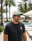 A man smiling outdoors by a pool wearing a dark charcoal t-shirt with a small white palm tree logo on the chest and a cream and green baseball cap with 'THINGS OFTEN Miami' printed on the front.