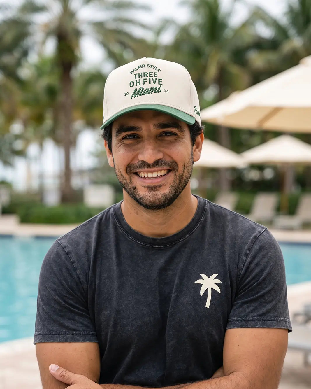 A smiling man wearing a dark charcoal t-shirt with a white palm tree logo and a cream baseball cap with green text, posed outdoors in front of a swimming pool with palm trees and umbrellas in the background.