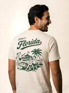 A man shown from the back wearing a cream-colored t-shirt with a green graphic print that reads 'Welcome to Florida' and features a retro illustration of a beach scene with palm trees, a house, and an alligator.