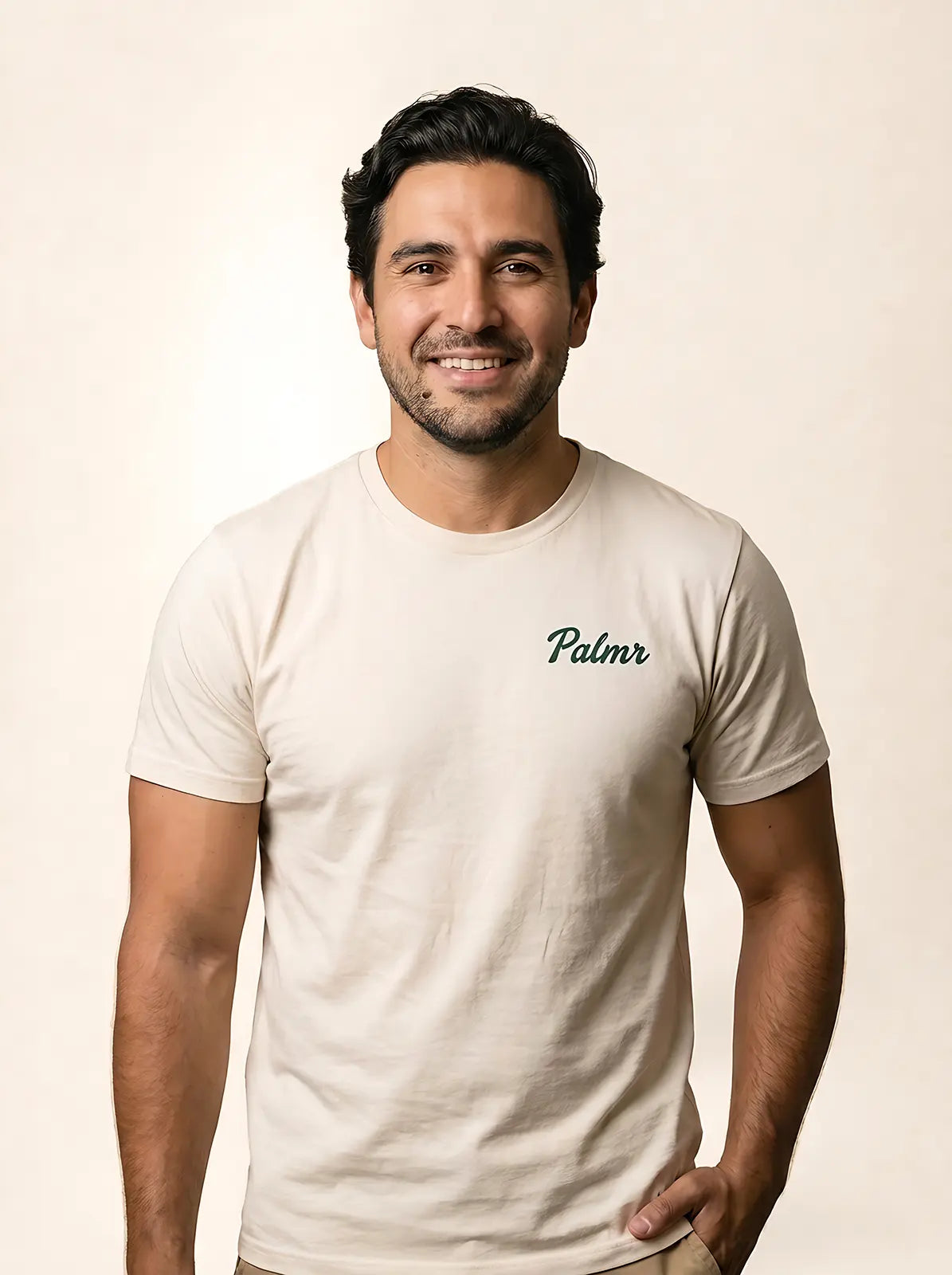 A smiling man wearing a light beige crewneck t-shirt with the brand name 'Palmr' printed in dark green script on the left chest, set against a plain beige background.